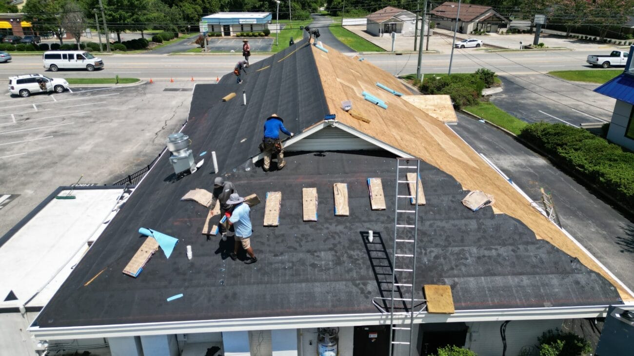 Aerial view of workers repairing a large commercial roof, some removing old shingles and others installing new roofing materials, with tools and ladders visible.