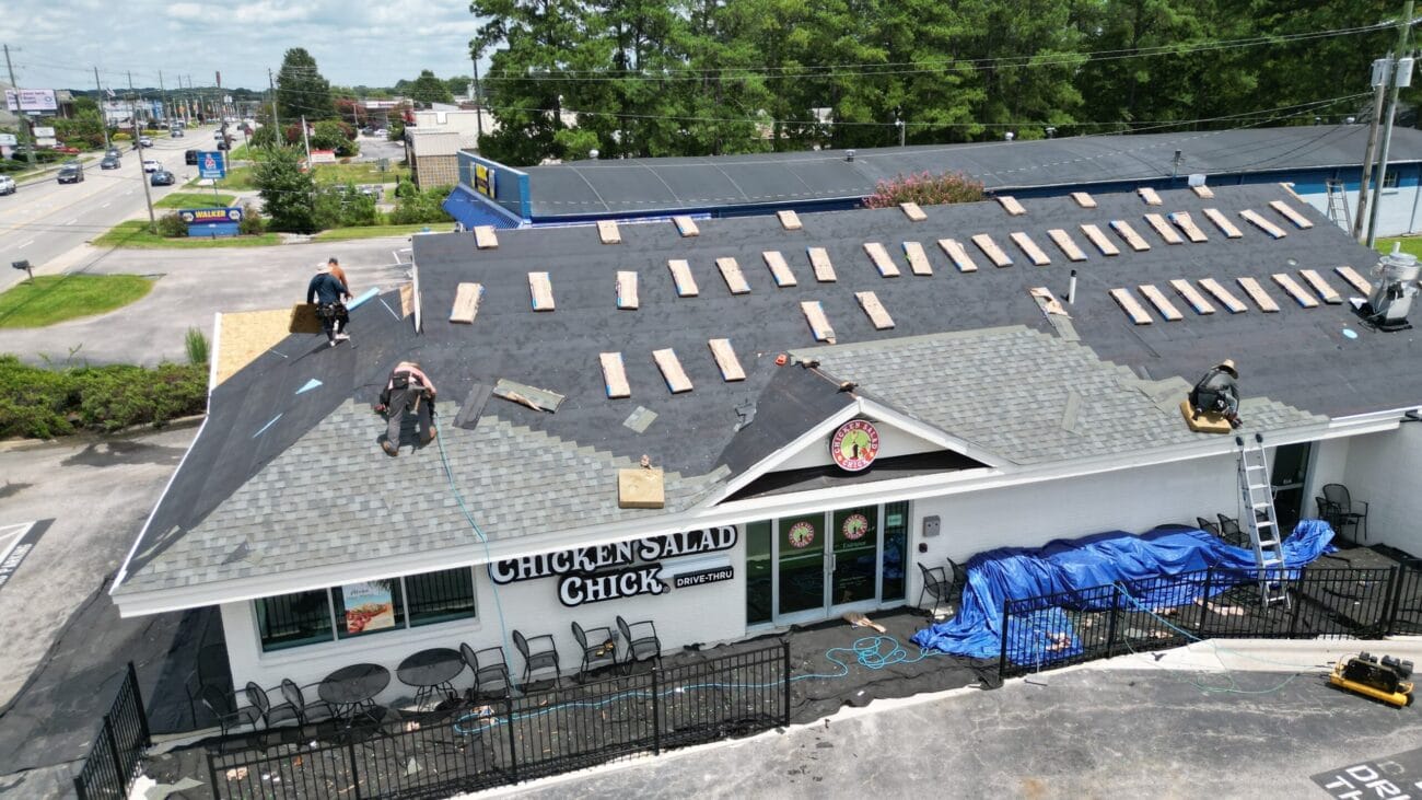 Workers are replacing shingles on the roof of a Chicken Salad Chick restaurant, with tools and materials spread out and protective tarps covering parts of the building and ground.
