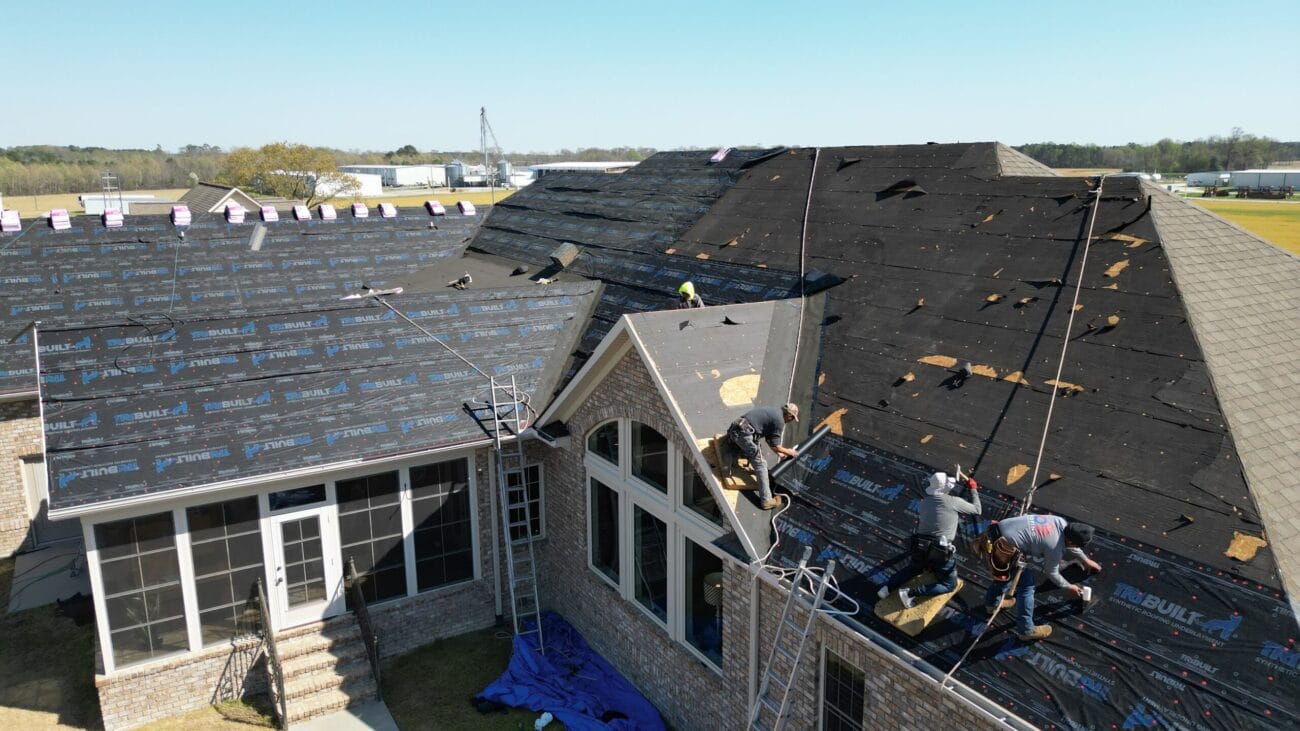 Several workers are installing new roofing materials on a large house; roofing tools, ladders, tarps, and safety ropes are visible on the roof and around the house.