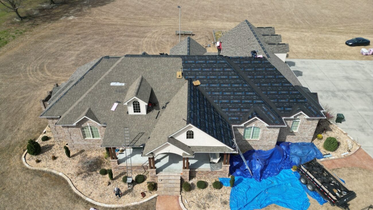 Aerial view of a house undergoing roof replacement, with workers, roofing materials, blue tarps, and part of the roof covered in underlayment.