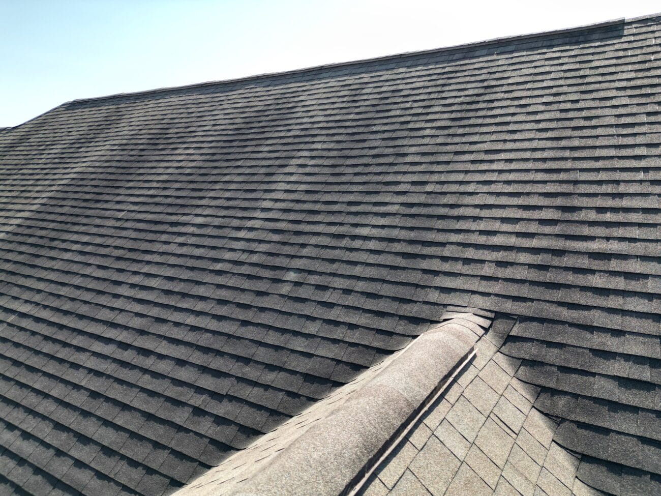 A close-up view of an asphalt shingle roof with overlapping rows and a ridge, under clear daylight.