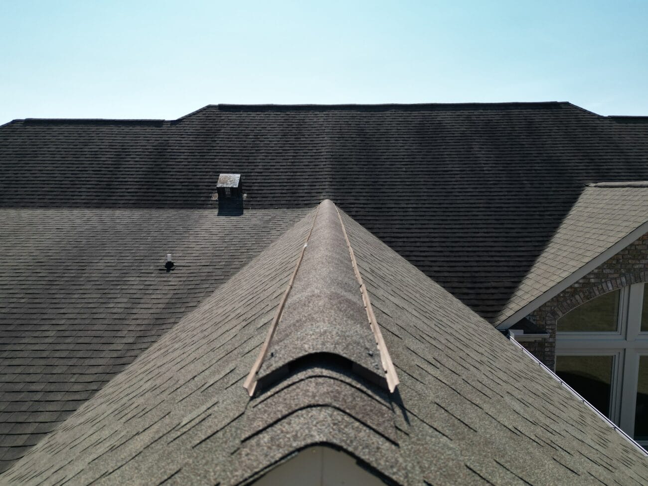 View of a residential roof ridge with asphalt shingles, showing a rooftop vent and parts of the house's sloped rooflines under a clear sky.