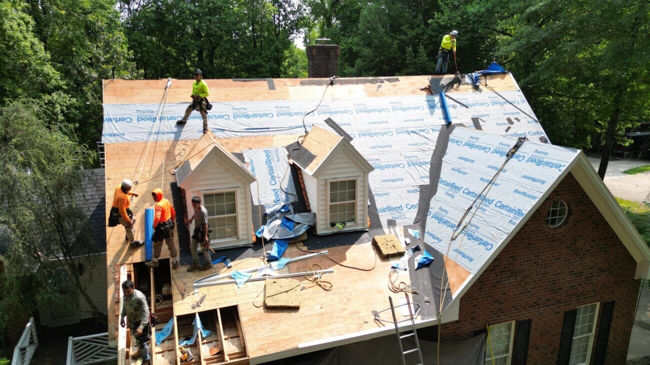 Several workers are installing roofing underlayment and materials on a partially stripped house roof, using safety harnesses and equipment.