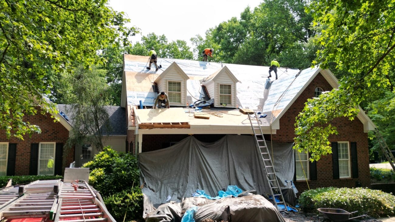 Workers are installing new roofing material on a house, with equipment and tarps covering the area below.