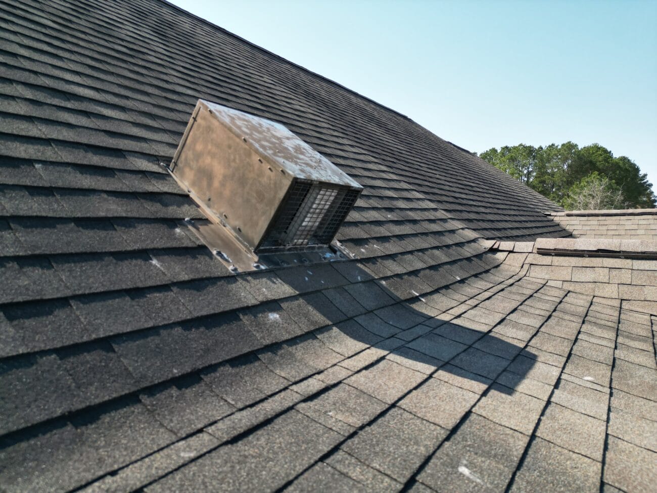A metal roof vent is installed on an asphalt shingle roof, with trees and clear sky in the background.