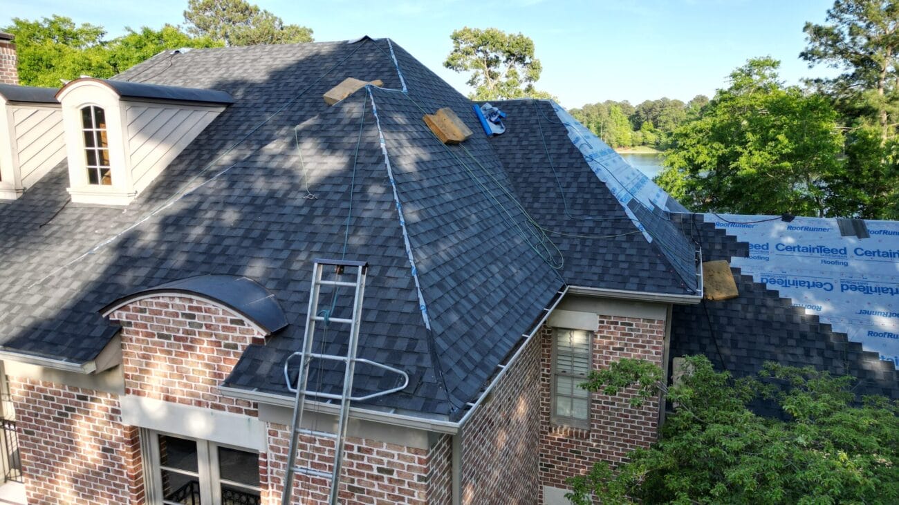 A brick house with dark gray shingle roofing under construction; some shingles and roofing materials are stacked on the roof, with a ladder leaning against the side.